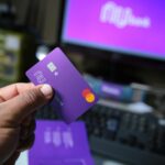 Close-up of a hand holding a purple bank card with a computer setup in the background, signifying online transactions.