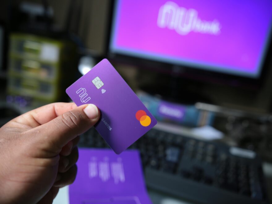 Close-up of a hand holding a purple bank card with a computer setup in the background, signifying online transactions.