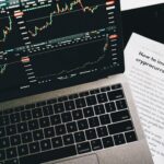 Close-up of a laptop displaying stock market graphs with a paper on investing in cryptocurrency, indoors.