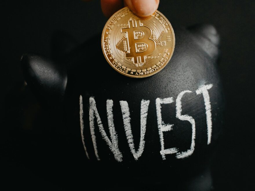 Gold bitcoin inserted into a black piggy bank labeled 'Invest', symbolizing cryptocurrency saving.
