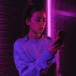 Young woman texting on mobile phone in a dark room with ambient pink lighting and a blurred background.
