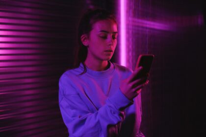 Young woman texting on mobile phone in a dark room with ambient pink lighting and a blurred background.