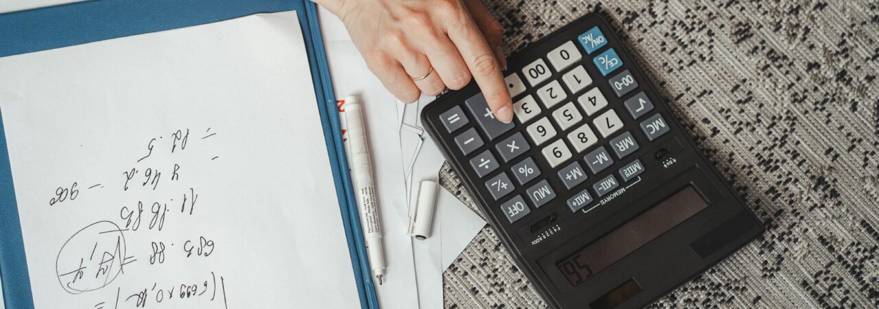 Top view of a hand calculating numbers with a large calculator and notes on a desk.