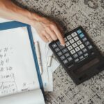 Top view of a hand calculating numbers with a large calculator and notes on a desk.