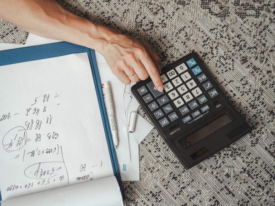 Top view of a hand calculating numbers with a large calculator and notes on a desk.